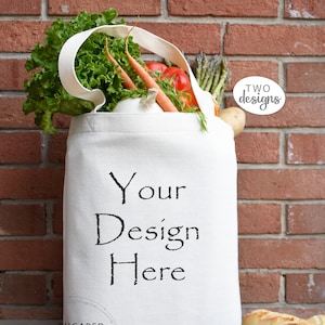 May include: White canvas tote bag with the text "Your Design Here" printed on the front. The bag is filled with fresh produce, including lettuce, carrots, and asparagus. The bag is sitting on a wooden surface.