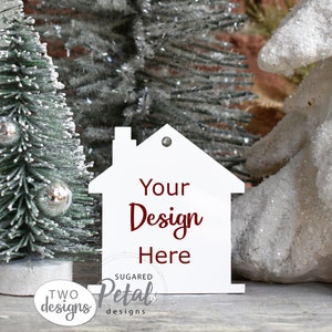 Puede incluir: Adorno de madera blanco en forma de casa con el texto rojo "Your Design Here" y un pequeño agujero en la parte superior para colgar. El adorno está sobre una superficie de madera con un pequeño árbol de Navidad artificial y un objeto decorativo blanco en el fondo.