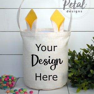Pode incluir: Uma cesta de Páscoa bege com uma alça e detalhes de orelhas de coelho. A cesta tem o texto "Your Design Here" impresso na frente. Uma pequena tigela de doces coloridos está à esquerda da cesta.