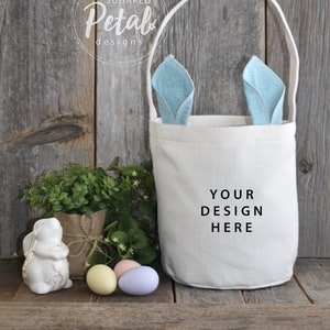 May include: A white canvas Easter basket with blue bunny ears. The basket has a handle and says "Your Design Here" on the front. The basket is sitting on a wooden surface with three Easter eggs and a white bunny figurine.