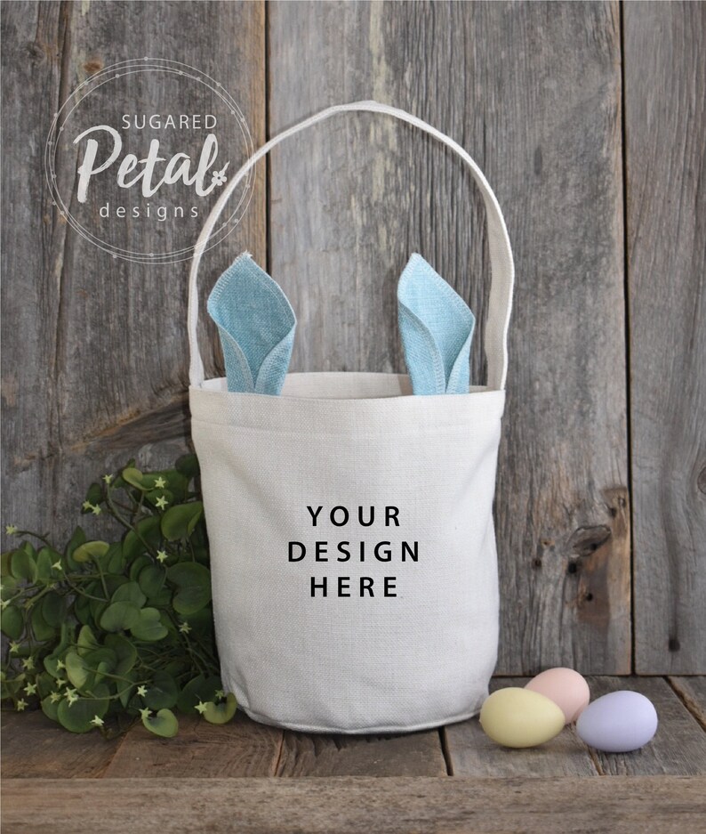 May include: A white canvas Easter basket with bunny ears made of blue fabric. The basket has a handle and says "YOUR DESIGN HERE" on the front. There are three pastel colored eggs in front of the basket.