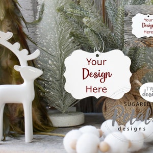 Op de afbeelding: Witte keramische rendierfiguur met een witte, sierlijke, lege kerstversiering met de tekst "Your Design Here" in rood. De versiering is omringd door witte kunstdennentakken, een in jute verpakt cadeau en witte wattenbolletjes.