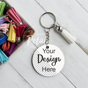 May include: A white round keychain with the text "Your Design Here" in black. The keychain has a silver chain and a white tassel. A bowl of colourful tassels is in the background.