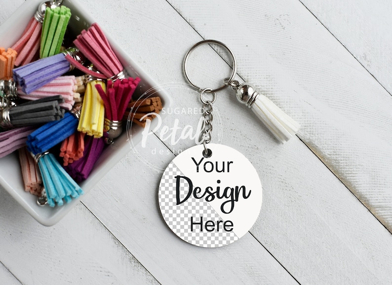 May include: A white round keychain with the text "Your Design Here" in black. The keychain has a silver chain and a white tassel. A bowl of colorful tassels is in the background.