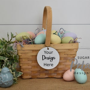 Op de afbeelding: Een rieten mand gevuld met pastelkleurige paaseieren, omringd door groene klimop en een blauw vogeltje. Een wit rond label met de tekst "Your Design Here" is aan de mand bevestigd.