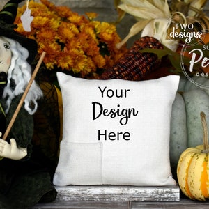 Peut inclure: Un oreiller blanc avec une poche et le texte "Your Design Here" sur une surface en bois rustique avec des décorations d'automne, y compris une poupée de sorcière, des citrouilles et du maïs.