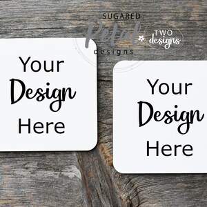 Puede incluir: Dos posavasos cuadrados blancos con el texto "Your Design Here" en fuente cursiva negra sobre un fondo de madera.