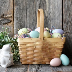 May include: A wicker basket filled with pastel coloured Easter eggs, sitting on a wooden surface. There is a small white ceramic bunny figurine next to the basket.