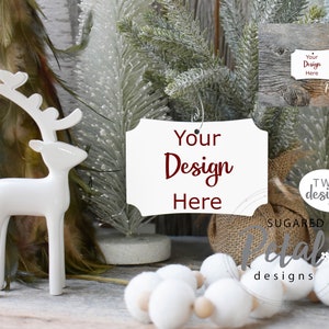 May include: White ceramic reindeer figurine with a blank white sign that says "Your Design Here" in maroon text. The sign is surrounded by white faux snow-covered pine branches and white pom poms.
