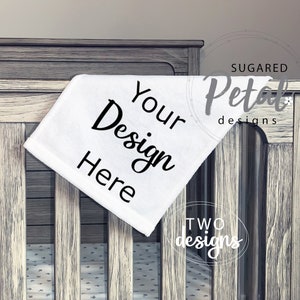 以下が含まれることがあります： 「Your Design Here」の文字が黒い筆記体で書かれた白いベビーブランケットが、灰色のベビーベッドに掛けられています。ベビーベッドには垂直の木製スラットがあります。ブランケットは柔らかく、カスタマイズ可能なアイテムです。