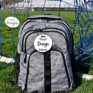 May include: A gray and black backpack with a white circle tag that says "Your Design Here" hanging from the zipper. The backpack is sitting on green grass in front of a blue soccer goal.