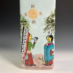 Pode incluir: Vaso retangular de cerâmica com uma cena nativa americana pintada à mão. A obra retrata duas figuras, um sol e árvores sobre um fundo azul claro. As figuras usam roupas coloridas. O vaso tem aproximadamente 20 cm de altura.