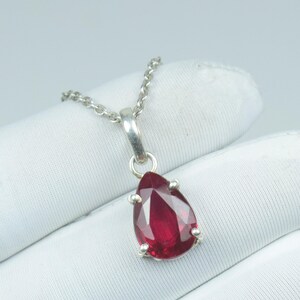 Peut inclure: Pendentif en argent avec une pierre précieuse rouge foncé en forme de goutte d'eau. La pierre est taillée à facettes et sertie dans un sertissage en argent. La chaîne est fine et en argent. Le bijou est présenté sur un fond blanc.