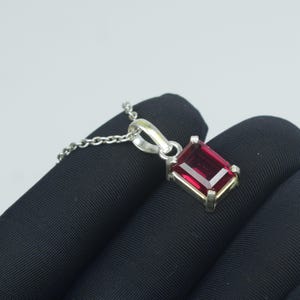 Peut inclure: Un collier pendentif en argent avec une pierre précieuse rectangulaire rouge foncé. La pierre est sertie dans un cadre argenté et suspendue à une chaîne délicate. Le pendentif est présenté sur un fond sombre.