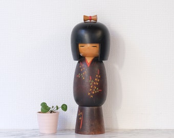 Puede incluir: Muñeca Kokeshi de madera con cabeza negra y cuerpo marrón. La muñeca tiene los ojos cerrados y un pequeño lazo rojo. El cuerpo está decorado con motivos florales amarillos, naranjas y verdes. Se ve una pequeña maceta rosa.