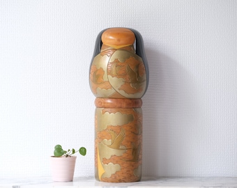 May include: A tall, wooden Japanese Kokeshi doll with a black head and a light brown face. The body is decorated with gold and brown designs of birds and clouds. A small pink pot with a green plant sits nearby.