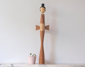 Könnte beinhalten: Eine Holzpuppe mit braunem Körper und einem schwarzen Haarknoten. Die Puppe hat ein weißes Gesicht mit schwarzen Augen und einem roten Mund. Die Puppe trägt ein braunes Kleid mit einem weißen Gürtel und einer braunen Schleife. Die Puppe steht auf einer weißen Oberfläche.
