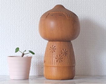 May include: A wooden doll with a rounded head and body, featuring carved details and a natural wood finish. The doll has a simple face and is decorated with star-like patterns. A small pink pot with a green plant is next to the doll.