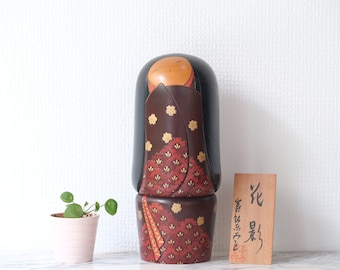 May include: A traditional Japanese wooden doll with a black and red patterned kimono. The doll has a round face with a painted expression and black hair. The doll is standing on a wooden base with a small sign that reads "花咲".
