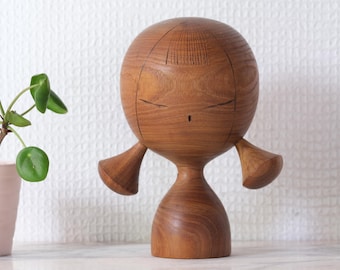 Könnte beinhalten: Holzfigur einer stilisierten Person mit rundem Kopf, geschlossenen Augen und kleinen Armen. Die Figur ist aus hellbraunem Holz gefertigt und hat eine glatte Oberfläche. Die Figur steht auf einer weißen Oberfläche.