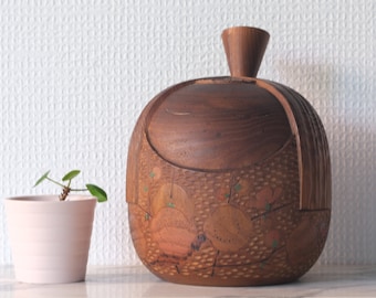 Puede incluir: Una caja de madera con un diseño tallado de flores y hojas. La caja es marrón y tiene una tapa con un pomo en la parte superior. La caja está sobre una superficie blanca.