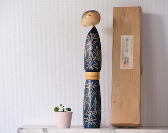 May include: A tall, dark blue Japanese Kokeshi doll with gold floral designs, topped with a small, light-colored head. A small potted plant and a brown box with Japanese text are also in the image. The doll is approximately 12 inches tall.
