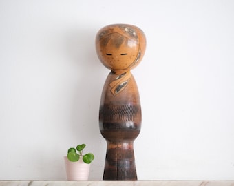 May include: A wooden kokeshi doll with a painted face and a patterned kimono. The doll is standing on a white surface with a small green plant in a pink pot.