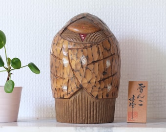 May include: A wooden sculpture of a figure with a rounded top and textured surface. The figure is light brown with darker brown accents and a small red detail. A small wooden sign with Japanese characters is next to the sculpture.