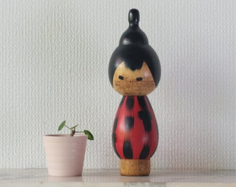 May include: A wooden doll with a black hair bun, red and black patterned dress, and a wooden base. The doll is standing on a white surface next to a small potted plant.