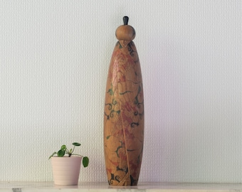 May include: A wooden decorative bottle stopper with a floral design in shades of red, green, and black. The stopper is shaped like a person and has a black knob on top.