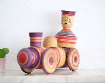 May include: A wooden toy train with a purple and yellow striped engine and a wooden figure with a striped hat and painted face. The figure is sitting on top of the engine.