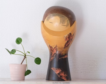 Op de afbeelding: Een hoog, glad houten beeldje met een afgeronde bovenkant en een taps toelopende voet. Het bovenste gedeelte is lichtbruin, terwijl het onderste gedeelte overgaat in een donkerbruine afwerking met gestructureerde, roodbruine bladvormige gravures.