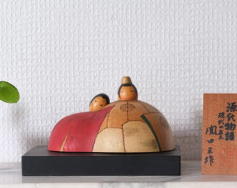 May include: A wooden Japanese doll with two figures, one larger and one smaller, on a tan and red base. The doll sits on a black stand. A wooden sign with Japanese characters is to the right.