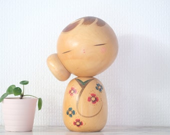 May include: A wooden Kokeshi doll with a rounded head and body, featuring painted details. The doll has closed eyes, rosy cheeks, and a hairstyle with brown accents. The body is adorned with floral patterns in red, blue, and green. A small pink pot with a green plant is next to the doll.