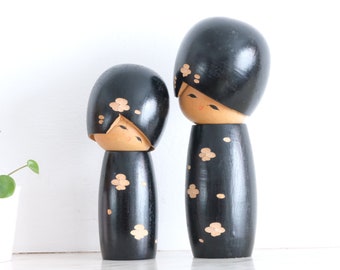 Puede incluir: Dos muñecas de madera negras con caras pintadas y diseños de flores blancas. Las muñecas llevan vestidos negros con diseños de flores blancas.