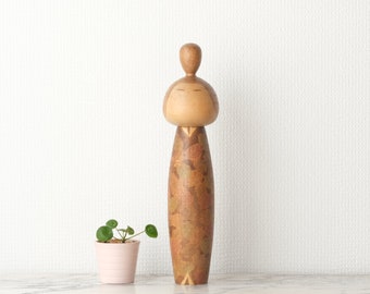 Könnte beinhalten: Eine japanische Holzpuppe mit einem braunen und goldenen gemusterten Kimono. Die Puppe hat ein einfaches Gesicht mit geschlossenen Augen und einen kleinen Knoten auf dem Kopf.