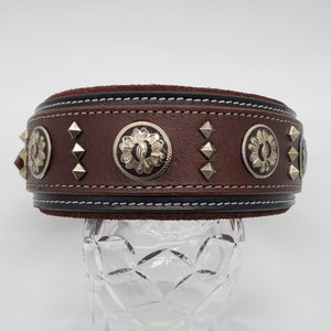May include: A brown leather dog collar with silver metal accents. The collar has a decorative stitching detail and is studded with silver pyramid-shaped studs.