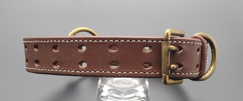 Puede incluir: Collar de perro de cuero marr&oacute;n con costuras blancas y herrajes de lat&oacute;n. El collar tiene varios agujeros para ajustar el tama&ntilde;o.