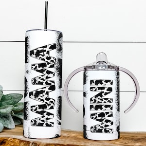 May include: A set of two stainless steel tumblers with a black and white cow print design. The larger tumbler has the word "MAMA" printed on it, and the smaller tumbler has the word "MINI" printed on it.