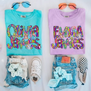 May include: Two turquoise and purple t-shirts with the names "Olivia James" and "Emma James" in colorful, patterned letters. The shirts are decorated with animal graphics, including tigers, penguins, and turtles.