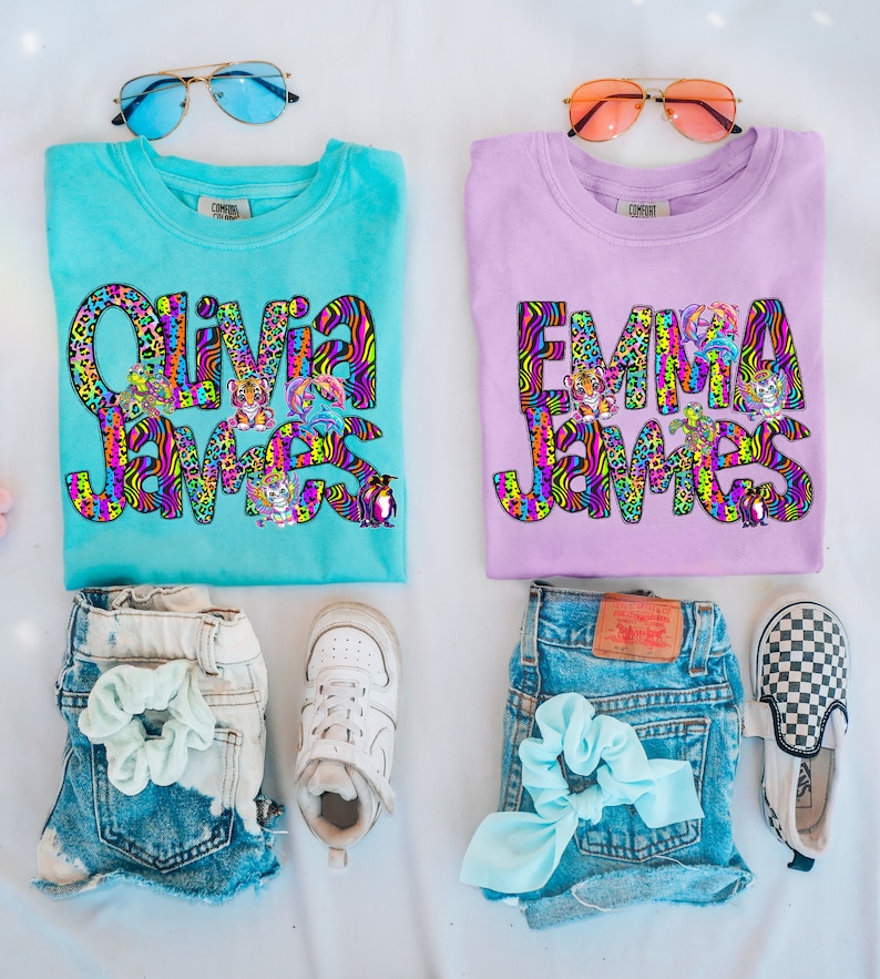 May include: Two turquoise and purple t-shirts with the names "Olivia James" and "Emma James" in colorful, patterned letters. The shirts are decorated with animal graphics, including tigers, penguins, and turtles.