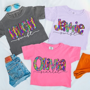 May include: Three colorful t-shirts with a name and last name printed on each. The names are Hugh, Jamie, and Olivia. The shirts are in different colors: gray, purple, and pink. The names are printed in a colorful, abstract pattern.