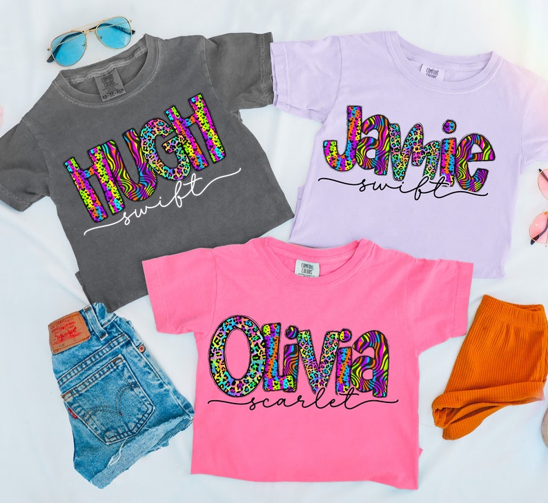 May include: Three colorful t-shirts with a name and last name printed on each. The names are Hugh, Jamie, and Olivia. The shirts are in different colors: gray, purple, and pink. The names are printed in a colorful, abstract pattern.