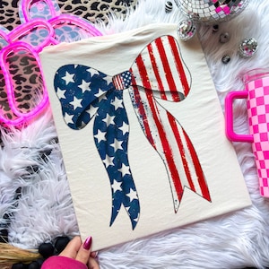 May include: A white t-shirt featuring a red, white and blue bow design. The bow is styled to resemble the American flag.
