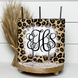 Puede incluir: Tres vasos altos transparentes con tapas y pajitas. Los vasos est&aacute;n decorados con un estampado de leopardo y un fondo blanco con un monograma negro "HS". Los vasos est&aacute;n colocados sobre un soporte de madera con un dise&ntilde;o decorativo.