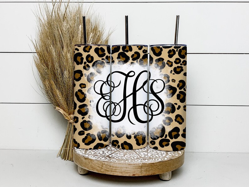 Puede incluir: Tres vasos altos transparentes con tapas y pajitas. Los vasos est&aacute;n decorados con un estampado de leopardo y un fondo blanco con un monograma negro "HS". Los vasos est&aacute;n colocados sobre un soporte de madera con un dise&ntilde;o decorativo.