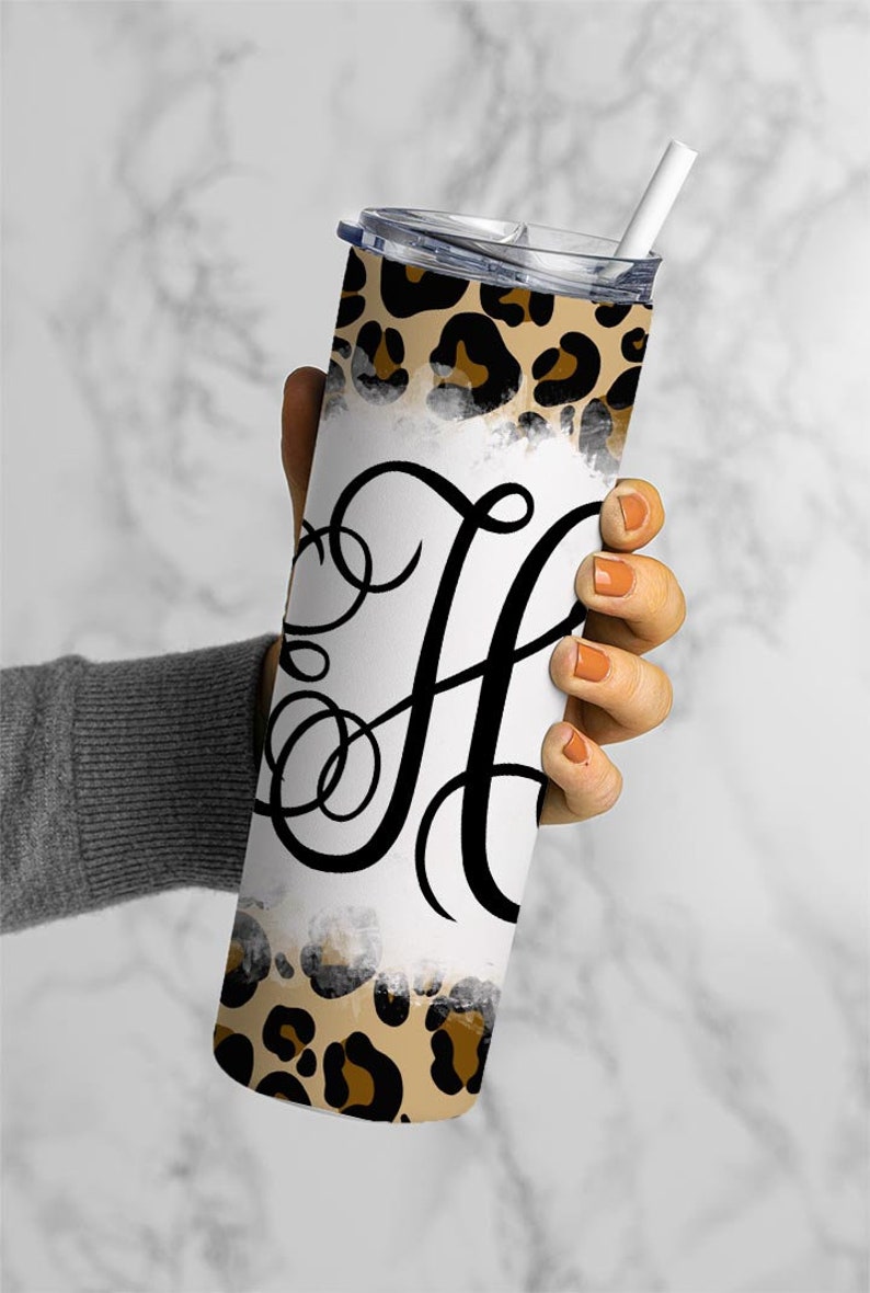 Puede incluir: Un vaso de acero inoxidable con fondo blanco y dise&ntilde;o de estampado de leopardo. El vaso tiene una letra "H" negra en cursiva en la parte delantera.