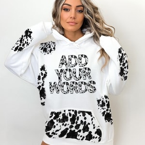 Puede incluir: Sudadera blanca con un diseño de estampado de vaca en blanco y negro. La sudadera tiene un gran gráfico negro que dice "ADD YOUR WORDS" en la parte delantera.