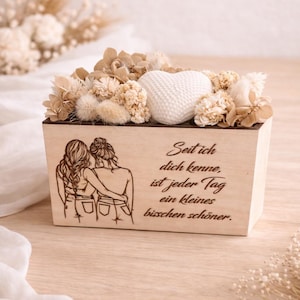 Puede incluir: Una caja de madera con una decoración en forma de corazón y flores secas. La caja tiene una ilustración grabada de dos personas abrazándose y texto en alemán. El arreglo está sobre una superficie de madera clara.