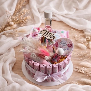 Könnte beinhalten: Ein herzförmiger Geschenkkorb mit rosa Schokoriegeln, einer kleinen Parfümflasche und Schönheitsprodukten. Der Korb enthält einen rosa Lippenbalsam, einen runden Behälter mit Balea Gesichtscreme und eine Flasche Disco Glitter Nagellack. Ein Holzschild trägt die Aufschrift "Mila".